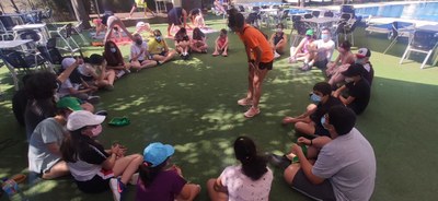 Grupo de chicos reunidos en corro atendiendo las explicaciones de la monitora mientras otros hacen estiramientos Grupo de chicos reunidos en corro atendiendo las explicaciones de la monitora mientras otros hacen estiramientos