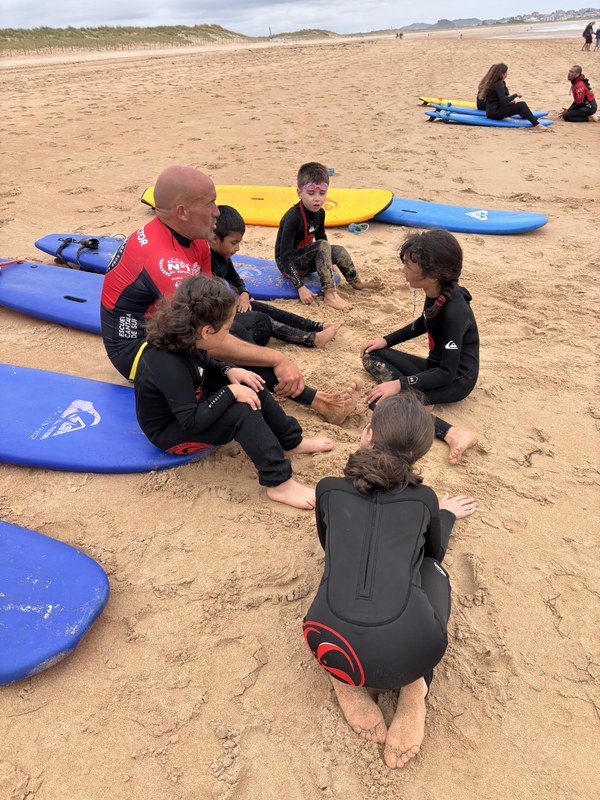 30.06 PEQUES PLAYA SURF.jpeg Participantes sentados junto al instructor y sus tablas de surf en la arena de la playa