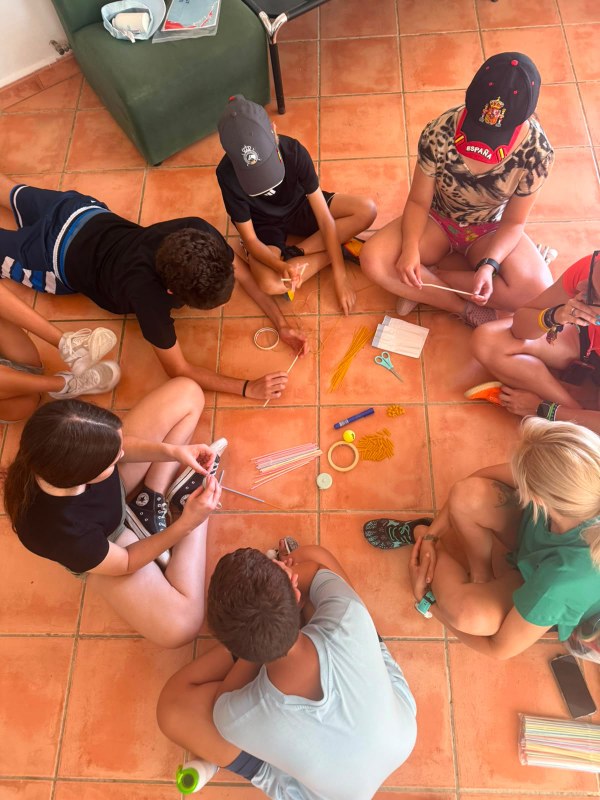 Grupo de participantes sentados en el suelo formando un círculo con los materiales en medio para la construcción de una estructura a partir de cinta, pajitas y espaguetis. y espa