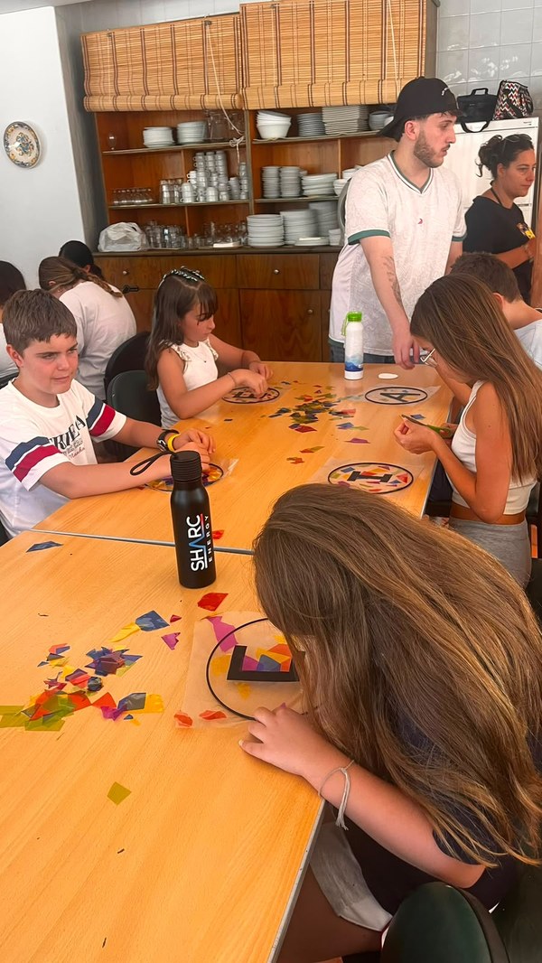Participantes sentados en una mesa trabajando en la decoración de la inicial de su nombre con papeles de colores simulando una vidriera