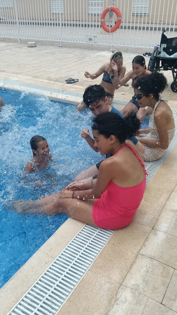 16.07.26 SALPICANDO EN LA PISCINA.jpeg Participantes sentado en la orilla de la piscina mientras los que están dentro del agua juegan a salpicarles.